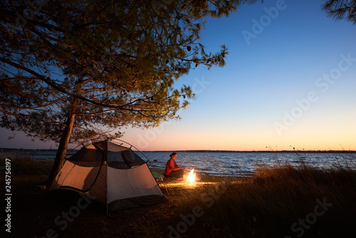 Camping on seasoast at night. Modern tourist tent under shadowy trees and hiker woman preparing food on campfire on blue evening sky and clear water background. Tourism and adventure concept.