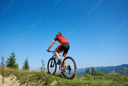 Back view of young athletic tourist biker in helmet and full equipment cycling bike up the grassy hill on distant mountains and blue summer sky background. Active lifestyle and extreme sport concept.