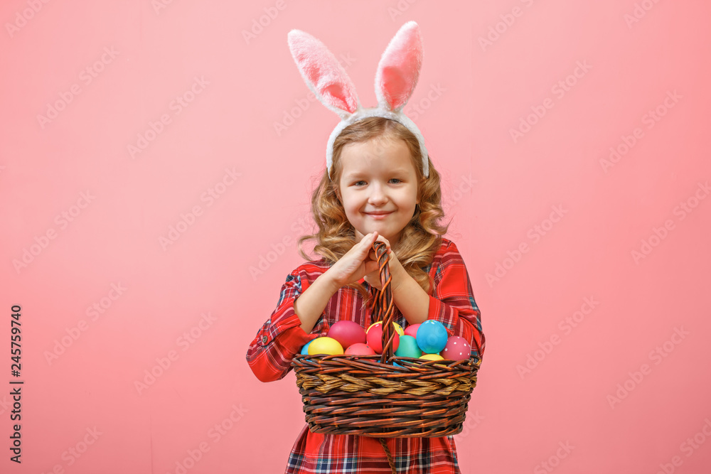 Cute little child girl with bunny ears holding basket of Easter eggs on a colored background.