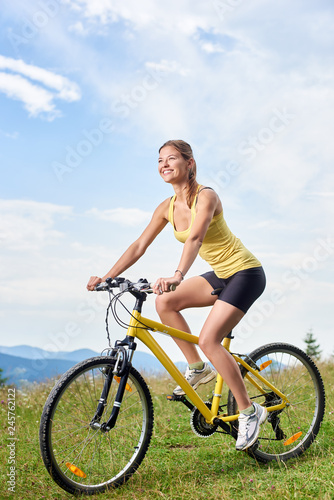 Vertical shot of attractive happy woman biker riding on yellow mountain bicycle, enjoying summer day in the mountains. Outdoor sport activity, lifestyle concept