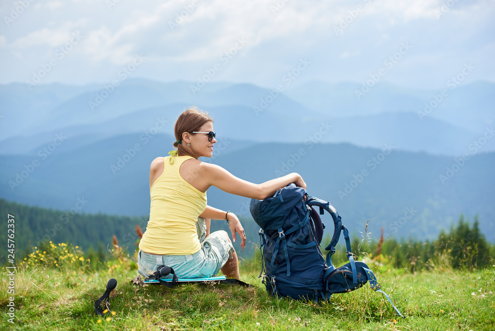 Back view of attractive woman tourist sitting on grassy hill with backpack, wearing sunglasses, enjoying summer cloudy day in the Carpathian mountains. Outdoor activity, tourism concept
