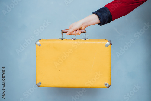 The hand of a girl in a red jacket holds a vintage yellow suitcase on a blue background