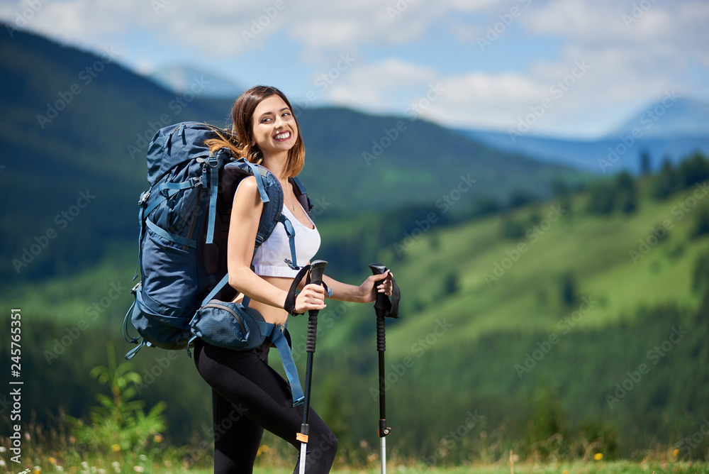 Portrait of attractive sporty female hiker with blue backpack and trekking poles, smiling on the top of a hill, enjoying summer day. Mountains, forests and cloudy sky on the blurred background