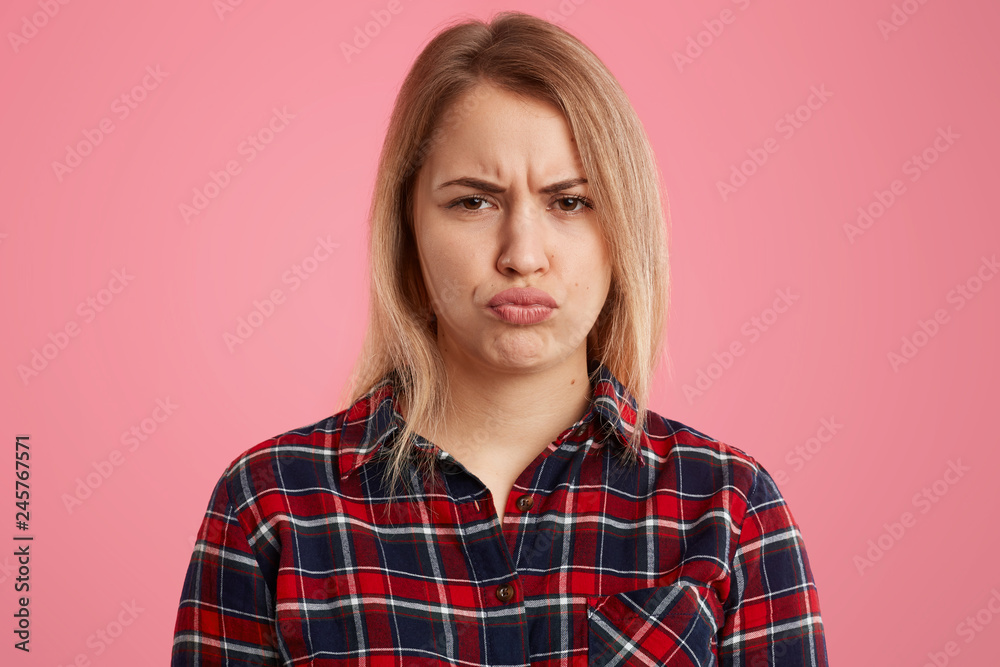 Indoor shot of displeased blonde woman pouts lips and frowns face, doesnt like something, has sullen facial expression, wears checkered shirt, isolated over pink background, makes dissatisfied grimace Indoor shot of displeased blonde woman pouts lips and frowns face, doesnt like something, has sullen facial expression, wears checkered shirt, isolated over pink background, makes dissatisfied grimace