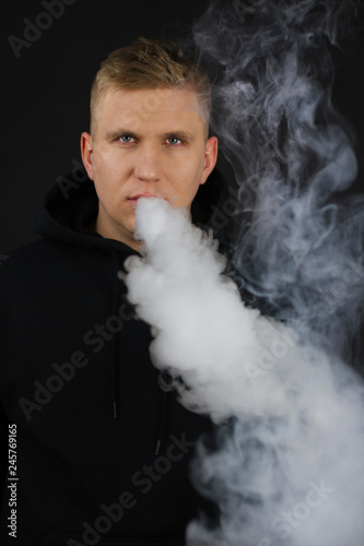 Man with vaping mod exhaling steam at black background. White guy smoking e-cigarette to quit tobacco. Vapor and alternative nicotine free smoking concept,blurred image