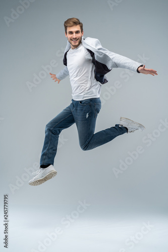 Full-length photo of funny man in casual t-shirt, blazer and jeans running or jumping in air isolated over gray background.