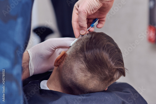 Barber is using razor while making a new hairstyle to a cute European boy. Side view.
