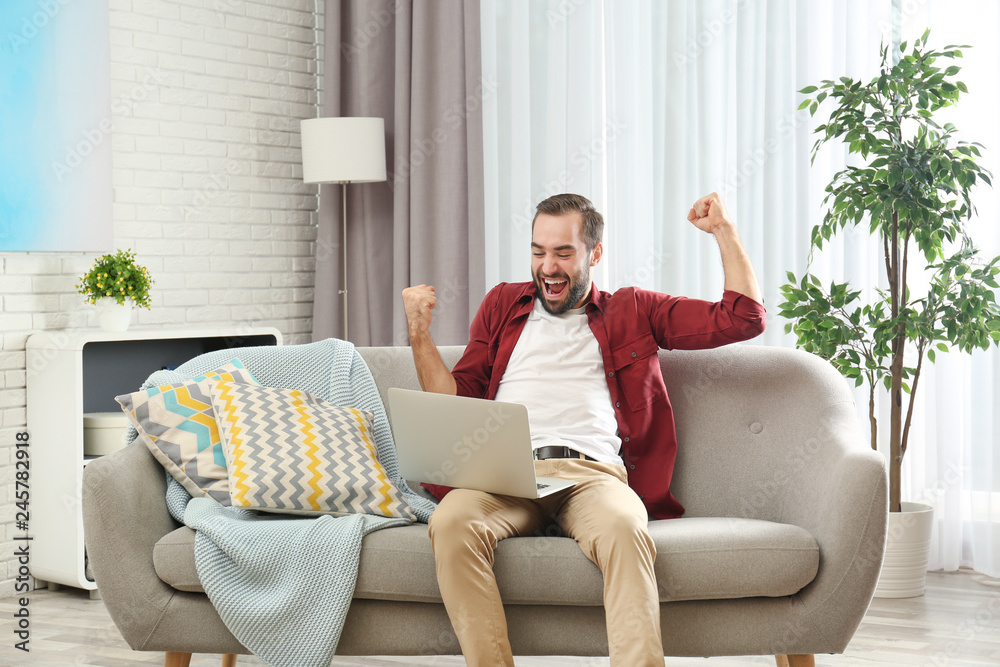 Obraz premium Emotional young man with laptop celebrating victory on sofa at home