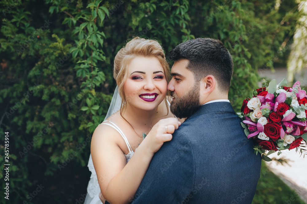 Stylish bearded groom and beautiful blonde bride hugging in a park on the nature. Portrait of lovers and smiling newlyweds in a green garden. Wedding photography. Close-up of newlyweds.