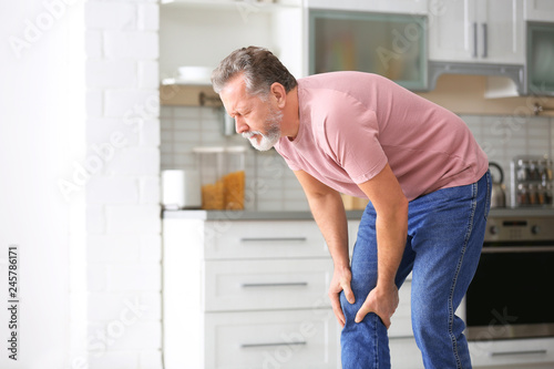 Senior man suffering from knee pain in kitchen. Space for text