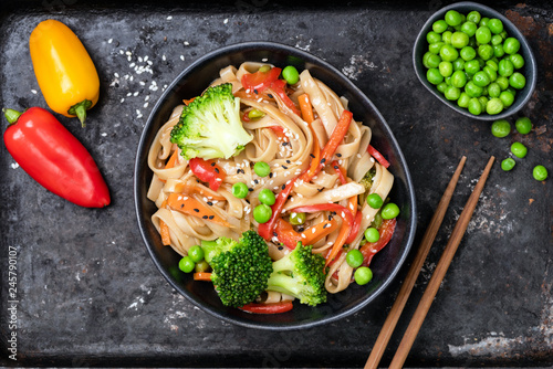 Vegetable stir fry with udon noodles, broccoli, carrot, pepper and green peas in bowl on black background. Top view photo