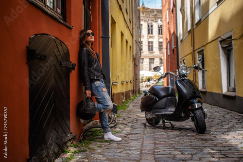 Fashionable girl holding a protective helmet while leaning on a wall, standing on an old narrow street.