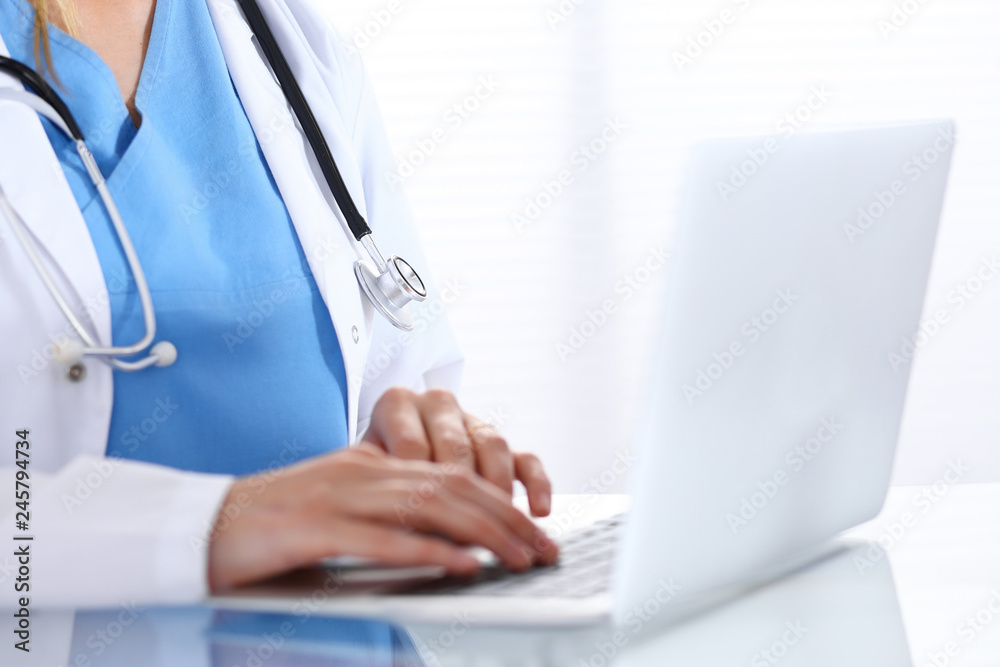 Doctor typing on laptop computer while sitting at the glass desk in hospital office. Physician at work. Medicine and healthcare concept Doctor typing on laptop computer while sitting at the glass desk in hospital office. Physician at work. Medicine and healthcare concept