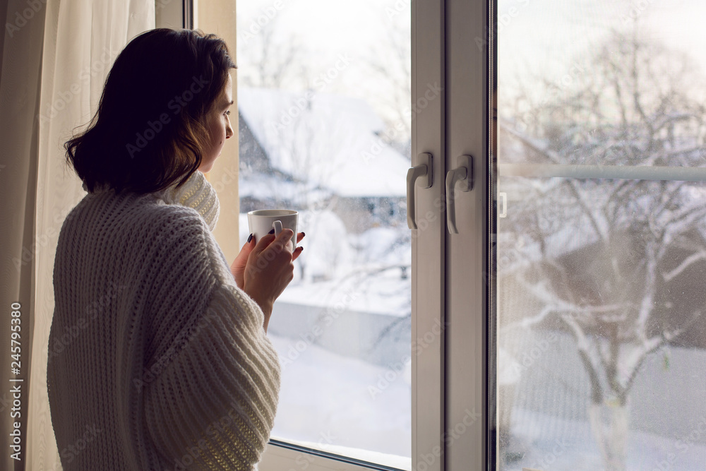 girl in a white knitted sweater stands at the window