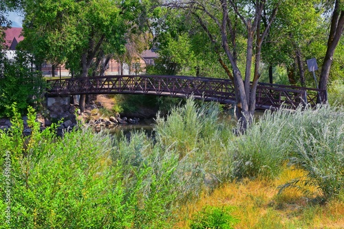 Views of Jordan River Trail Pedestrian and Train Track Bridge with surrounding trees, Russian Olive, cottonwood and muddy stream along the Wasatch Front Rocky Mountains, in Salt Lake City, Utah.