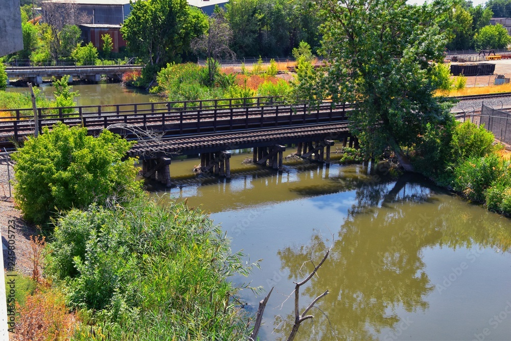 Fototapeta premium Views of Jordan River Trail Pedestrian and Train Track Bridge with surrounding trees, Russian Olive, cottonwood and muddy stream along the Wasatch Front Rocky Mountains, in Salt Lake City, Utah.