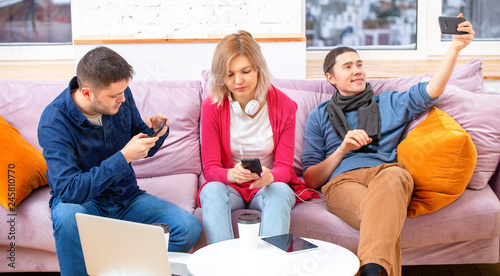 Friends sit on the couch, chatting and using a smartphone, desktop computer, laptop.