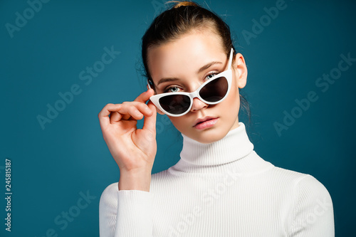 Portrait of a beautiful young girl in white knitted golf and sunglasses on a blue background