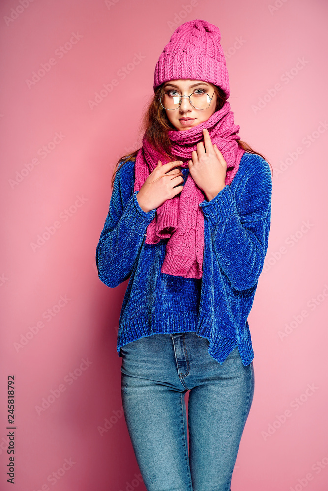 Modern young woman wearing blue sweater and pink hat, and scarf posing, making funny facial expression. Retouched, studio portrait. Modern young woman wearing blue sweater and pink hat, and scarf posing, making funny facial expression. Retouched, studio portrait.