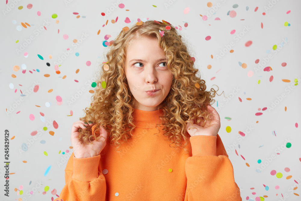Horizontal shot of attractive young female with pensive contemplating expression,ponders about something,keeps touches her blond hair,dressed in casual orange sweater, stands under falling confetti Horizontal shot of attractive young female with pensive contemplating expression,ponders about something,keeps touches her blond hair,dressed in casual orange sweater, stands under falling confetti