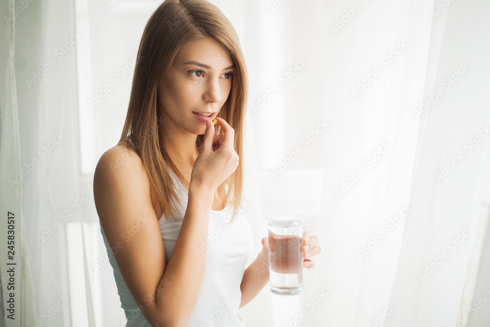 Happy smiling positive woman eating the pill and holding the glass of water in the hand, in her home - obrazy, fototapety, plakaty 