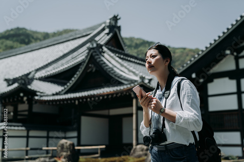 girl photographer holding cellphone checking the tourist route while self guided travel in kyoto japan. young asian female backpacker carrying camera searching direction on online map in Tenryu ji. photo