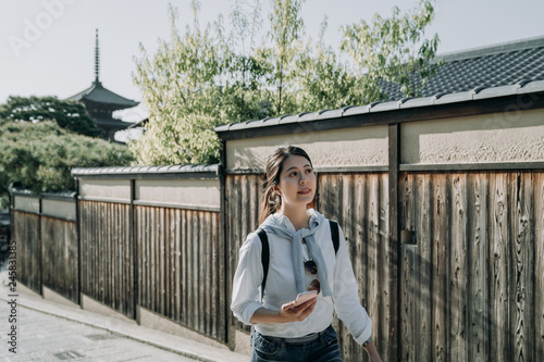 young woman backpacker holding smart phone searching direction to the next sightseeing spot after visit famous attraction Hokanji pagoda. girl traveler using online map app walking alone wood wall. photo