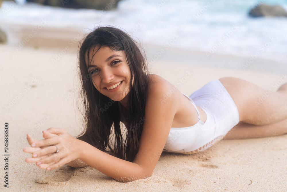 Portrait of beautiful smiling girl in white swimsuit lying on beach in waves of ocean