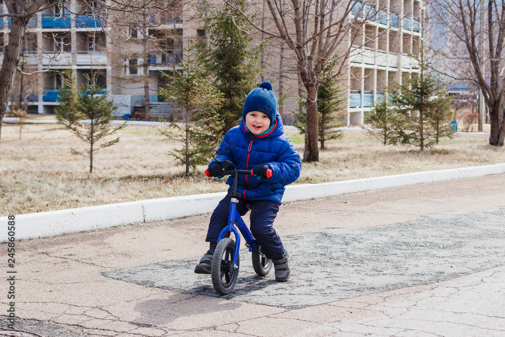cheerful little boy in warm clothes riding a children's running bike in early spring