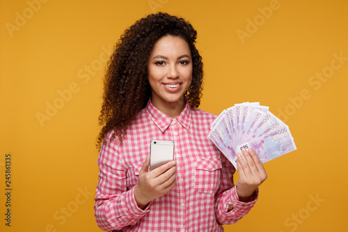 Portrait of an excited young african isolated over yellow background. Looking camera showing display of mobile phone holding money