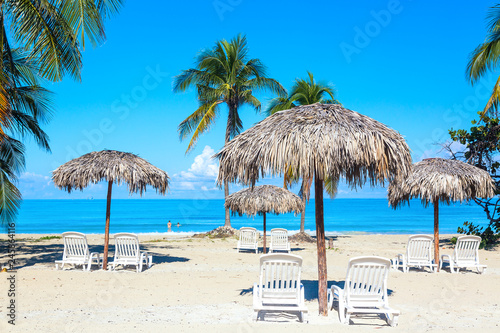 A sun loungers under umbrellas on the sandy beach with palms by the sea and sky. Vacation background. Idyllic beach landscape.