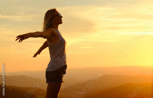 Happy woman with open arms stay on the peak of the mountain cliff edge under sunset light sky enjoying the success  freedom and bright future