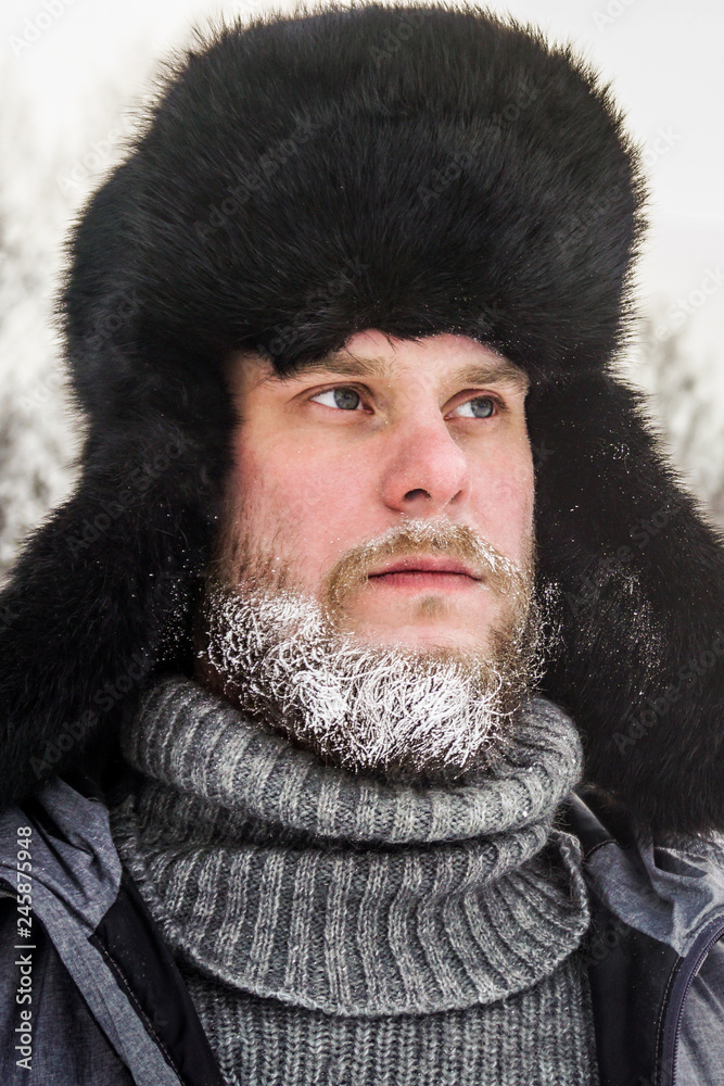 Siberian Russian man with a beard in hoarfrost in freezing cold in the winter freezes in a village in a snowdrift and wears a hat with a earflap. Siberian Russian man with a beard in hoarfrost in freezing cold in the winter freezes in a village in a snowdrift and wears a hat with a earflap.