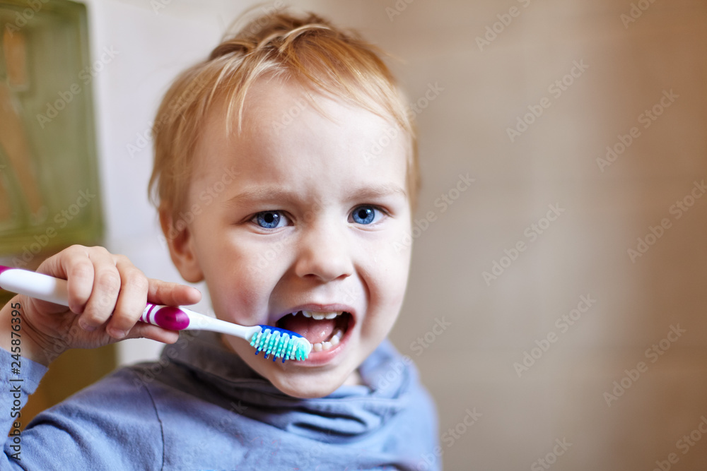 Close up portrait of cute caucasian baby boy with very serious face expression trying to clean the teeth with teeth brush, by himself. Bright blue eyes, fair hair. Indoors, copy space. Close up portrait of cute caucasian baby boy with very serious face expression trying to clean the teeth with teeth brush, by himself. Bright blue eyes, fair hair. Indoors, copy space.