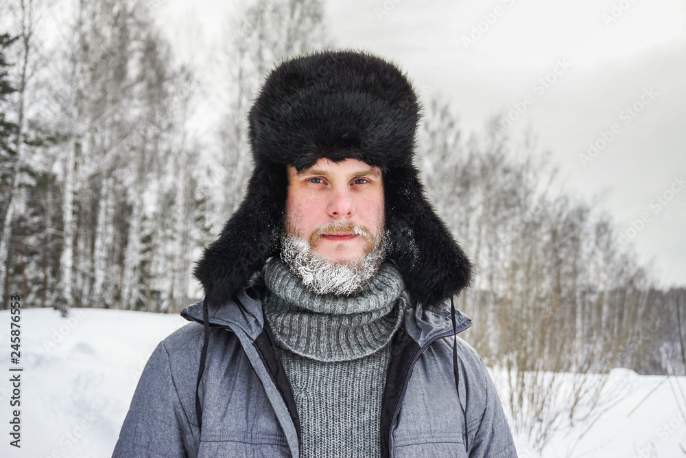 Siberian Russian man with a beard in hoarfrost in freezing cold in the winter freezes in a village in a snowdrift and wears a hat with a earflap. Siberian Russian man with a beard in hoarfrost in freezing cold in the winter freezes in a village in a snowdrift and wears a hat with a earflap.