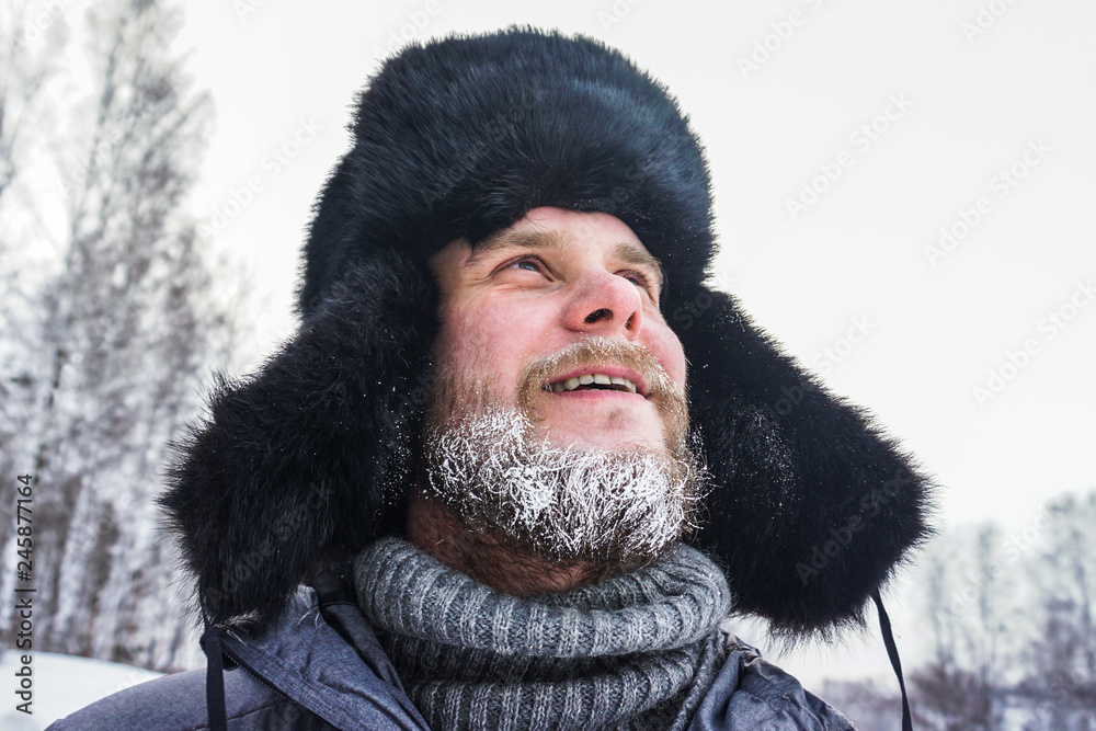 Happy Siberian Russian man with a beard smiles and lets out steam from his mouth in hoarfrost in freezing cold in the winter freezes in a village in a snowdrift and wears a hat with a earflap. Happy Siberian Russian man with a beard smiles and lets out steam from his mouth in hoarfrost in freezing cold in the winter freezes in a village in a snowdrift and wears a hat with a earflap.