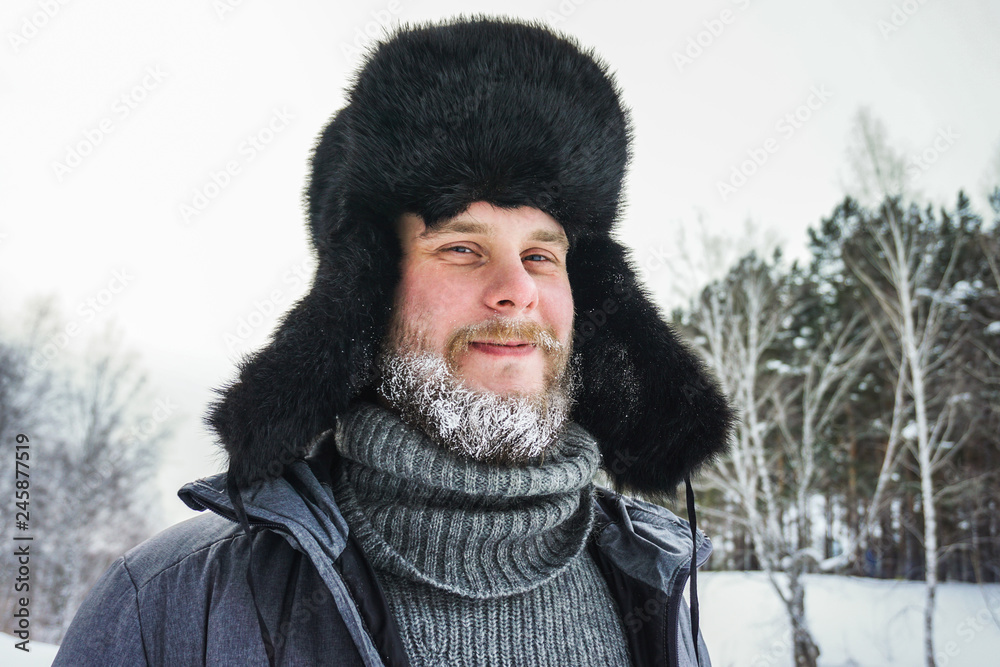Happy Siberian Russian man with a beard smiles and lets out steam from his mouth in hoarfrost in freezing cold in the winter freezes in a village in a snowdrift and wears a hat with a earflap. Happy Siberian Russian man with a beard smiles and lets out steam from his mouth in hoarfrost in freezing cold in the winter freezes in a village in a snowdrift and wears a hat with a earflap.