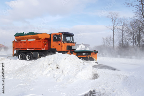 A large car with a plow clears the road from snow. Orange cargo special equipment is struggling with the elements in winter. Removing the effects of the snowstorm. Difficulties in traffic.Frozen water