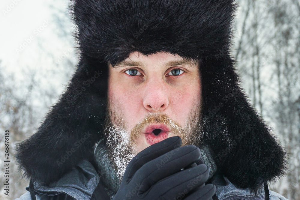 a frozen man with a beard with weathered skin and lips in the winter in a forest with snowdrifts warms his hands with the cold from a walk after walking through the village a frozen man with a beard with weathered skin and lips in the winter in a forest with snowdrifts warms his hands with the cold from a walk after walking through the village