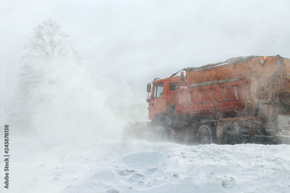 A large car with a plow clears the road from snow. Orange cargo special equipment is struggling with the elements in winter. Removing the effects of the snowstorm. Difficulties in traffic.Frozen water A large car with a plow clears the road from snow. Orange cargo special equipment is struggling with the elements in winter. Removing the effects of the snowstorm. Difficulties in traffic.Frozen water