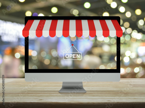 Desktop modern computer monitor with online shopping store graphic and open sign on wooden table over blur light and shadow of shopping mall, Business internet shop online concept