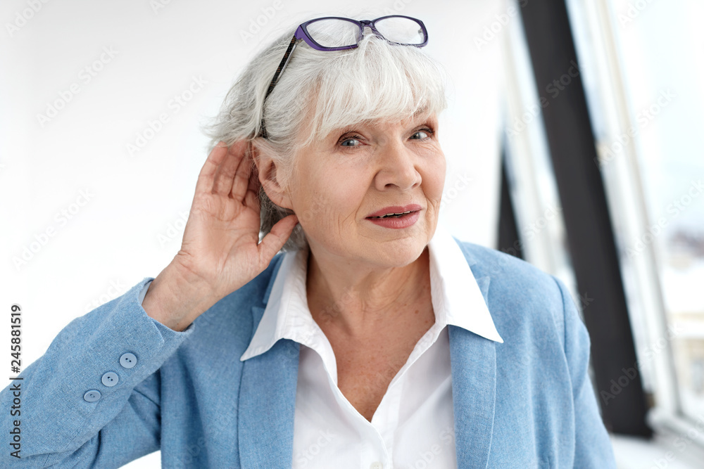 Close up elegant stylish elderly woman in formal suit having hearing problems, holding hand at her ear, trying to hear you, saying: Speak louder, please. Age, maturity, people and health concept Close up elegant stylish elderly woman in formal suit having hearing problems, holding hand at her ear, trying to hear you, saying: Speak louder, please. Age, maturity, people and health concept