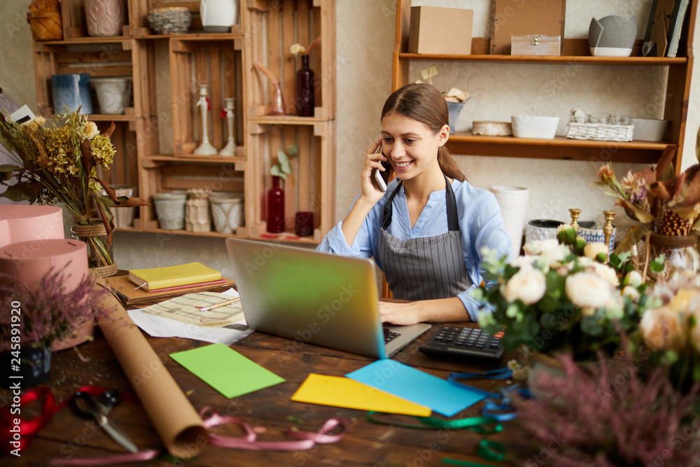 © Seventyfour - Portrait of female small business owner using laptop and speaking by phone in flower shop, copy space