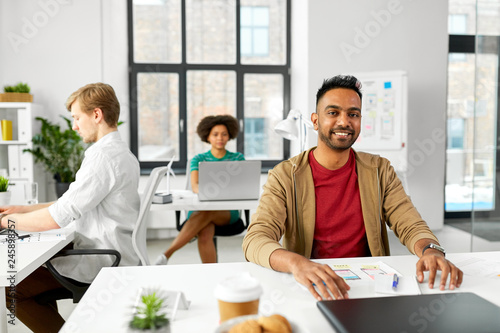 business and people concept - happy smiling indian male creative worker with laptop sitting at office table