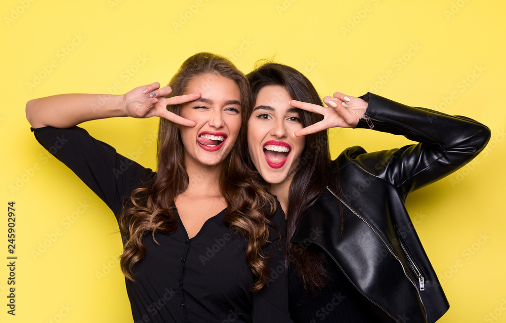 Two playful girls gesturing v-sign near eyes on yellow Two playful girls gesturing v-sign near eyes on yellow