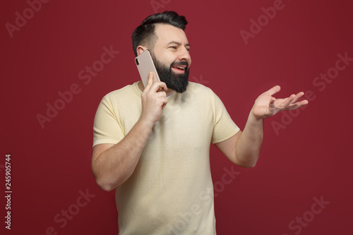 A cute and bearded brunette man on a red background says emotionally on the phone