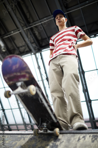 Teenage guy in striped tee and beige pants standing on edge of parkour area before descending on skateboard