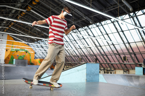 Young casual lad with vr headset standing on skateboard while moving along flat surface of parkour area
