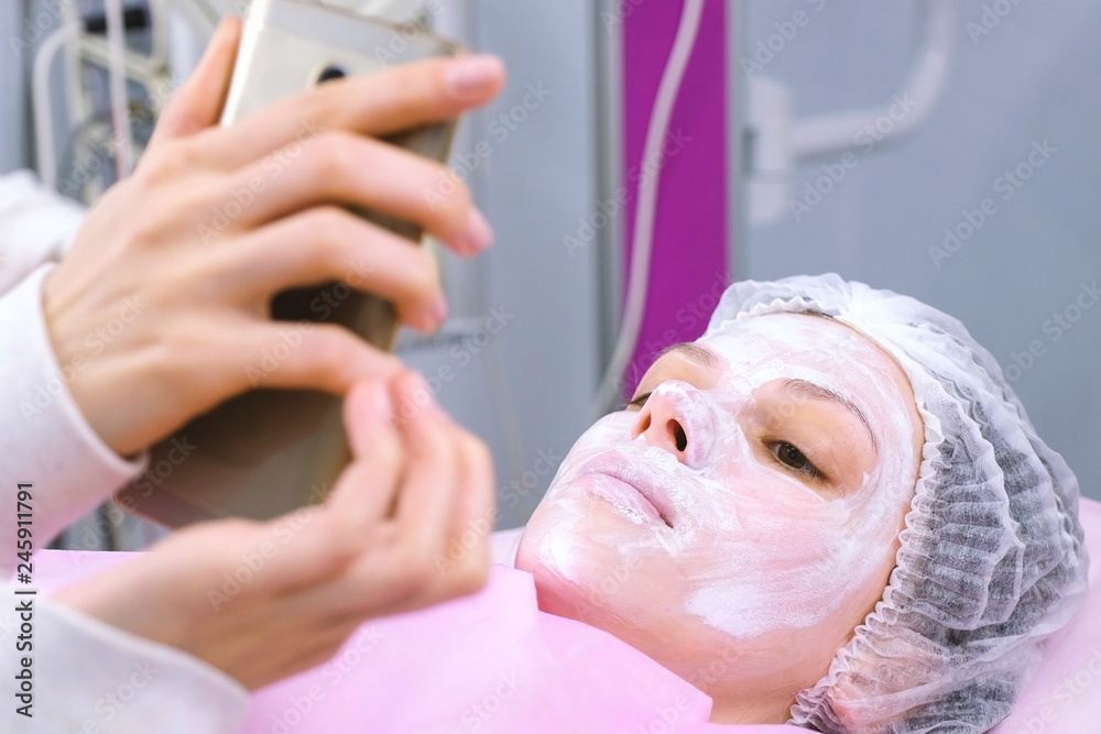Woman reads something in a mobile phone lying on the procedure in the beautician's office with a mask on her face.