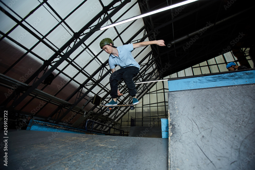 Boy in helmet and casualwear standing on board while moving in jump over skateboarding racetrack